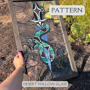 May include: A stained glass window featuring a green snake wrapped around a dagger. The snake has a brown and white pattern. The window is framed in wood and has the text "PATTERN" and "DESERT WILLOW GLASS" on it.