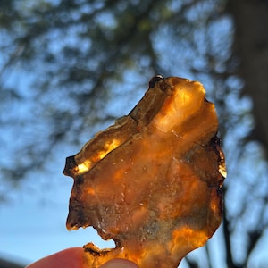 May include: A translucent, amber-colored stone held up against a blue sky. The stone has a rough, irregular shape with a mix of light and dark brown hues. The light shines through the stone, highlighting its unique texture.