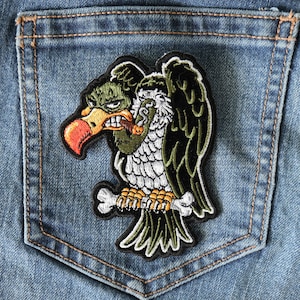 May include: A green, black, and white embroidered patch of a cartoon vulture with a cigarette in its beak, perched on a bone. The patch is sewn onto a blue denim pocket.