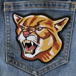 May include: A brown and white embroidered patch of a snarling cougar's head on a blue denim background.