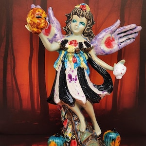 May include: A ceramic figurine of a woman with wings, holding a mask and a small white object. The figurine has a black and white striped dress, and is standing on a decorative base with two blue and orange spheres at the base.
