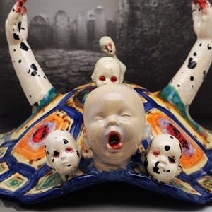 May include: A ceramic sculpture featuring a central figure with an open mouth, surrounded by baby doll heads. The figure has outstretched arms with red-painted fingertips and black spots. The base is a colorful, patterned design. A crow sits atop a tombstone in the background.