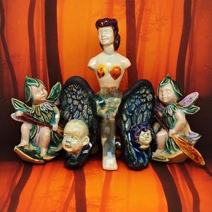 May include: A ceramic sculpture featuring a central figure with a woman's torso, angel wings, and multiple baby heads. Two cherubic figures with wings flank the central figure. The sculpture is painted in shades of green, blue, and brown.