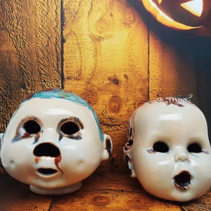May include: Two ceramic doll heads with painted details. One head has blue and white paint, the other has brown and white paint. Both heads have painted eyes, noses, and mouths.