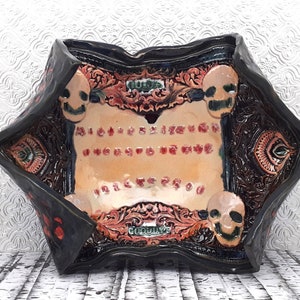 May include: A black ceramic bowl with a decorative design featuring skulls and floral patterns. The bowl has a raised rim and a flat bottom. The inside of the bowl is painted with a light brown glaze and features a red and white Ouija board design.