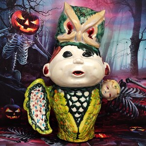May include: A ceramic figurine of a child with an owl head and wings. The figurine is painted in bright colors, including green, yellow, red, and blue. The child is wearing a green and yellow jacket with a black and white pattern. The figurine is set against a dark background with a spooky forest scene.