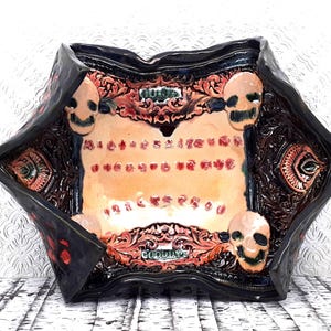 May include: A ceramic decorative bowl with a unique, gothic design. The bowl features a black exterior with red accents and a central tan area with red dots. Four smiling skulls are placed around the bowl's interior. The word "GOODBYE" is visible.