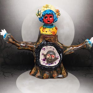 May include: A ceramic sculpture featuring a tree trunk with outstretched arms. The top has a doll-like face with red skin, blue hair, and a yellow hat. The trunk has a dark opening with a baby-like face inside. The sculpture is on a white base.
