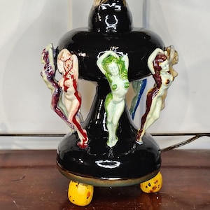 May include: A black ceramic lamp with a glossy finish, featuring multiple figurines of nude women in various poses. The lamp has a bulbous shape with yellow feet and a power cord.