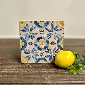 May include: A square wooden block with a blue and yellow floral tile design. A yellow lemon and green sprig sit to the right of the block on a wooden surface. The tile design features blue leaves and yellow flower centers.