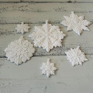 May include: A collection of six white snowflake ornaments featuring intricate, embossed floral and scrollwork patterns. Each piece has a small circular loop at the top for hanging, displaying a matte finish with detailed, symmetrical, and ornate designs.