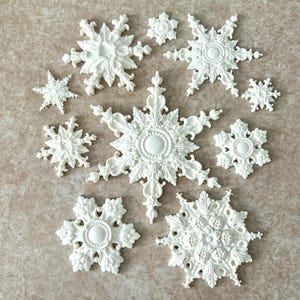 May include: A collection of ten off-white, molded decorative ornaments shaped like snowflakes. Each piece features intricate, raised relief patterns with floral and scrollwork details, varying in size and design, arranged in a cluster.
