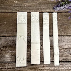 May include: Four white rectangular objects resembling book spines arranged vertically. Each piece features distinct embossed decorative patterns, including floral motifs, horizontal bands, and blank rectangular panels, all rendered in a matte white finish.