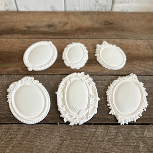 May include: Six white oval-shaped frames with varied decorative borders. The frames feature intricate relief details including floral patterns, beaded edges, and leafy vine motifs. Each frame has a smooth, solid white center surface for display.