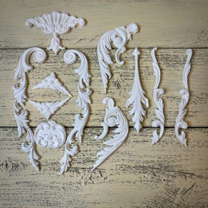 Peut inclure: Un ensemble d'appliques décoratives en résine blanche présentant des volutes complexes, des motifs de feuilles d'acanthe et des designs floraux symétriques. Ces pièces ornementales incluent des angles courbés, un cimier central et des moulures verticales.