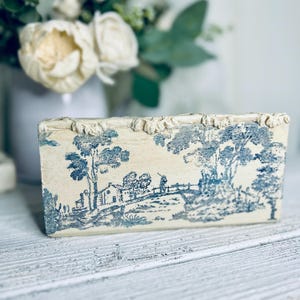 May include: A rectangular, off-white box with a blue toile landscape design. The box has a textured, decorative border along the top edge. The toile scene depicts trees, buildings, and figures, creating a classic, vintage aesthetic.