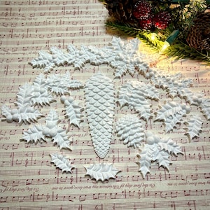 May include: White Christmas-themed decorative elements, including holly leaves, berries, and pine cones. The assortment is arranged on a musical score background, with a festive evergreen garland and lights in the corner.