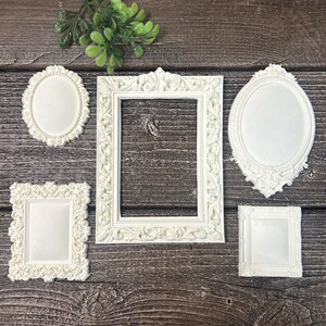 May include: A collection of five off-white picture frames featuring intricate raised scrollwork and floral relief patterns. The set includes one large rectangular frame, two smaller rectangular frames, one oval frame, and one large ornate oval frame.