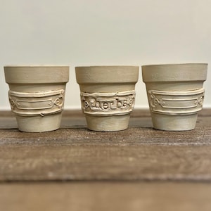 May include: Three off-white plant pots arranged in a row. Each pot features a raised rectangular decorative plaque on the front. The center pot has the word herbs embossed on its plaque, while the two outer pots feature ornate scrollwork.