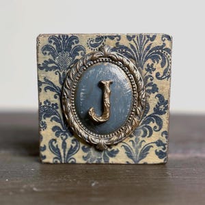 May include: A decorative wooden block featuring a blue and cream floral pattern. A distressed oval frame with a gold-colored trim surrounds a blue painted surface with the letter "J" in gold.