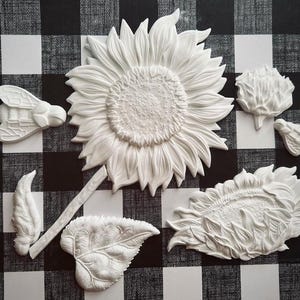 Puede incluir: Un conjunto de elementos decorativos blancos y texturizados con forma de girasoles y abejas. El conjunto incluye un girasol grande, un girasol más pequeño, una abeja, una hoja y otros componentes florales. Los elementos están dispuestos sobre un fondo a cuadros blanco y negro.
