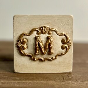 May include: A square, off-white block with a gold-colored, ornate frame. The frame surrounds a gold letter "M". The block is likely made of wood or a similar material, and the design has a vintage or antique aesthetic.