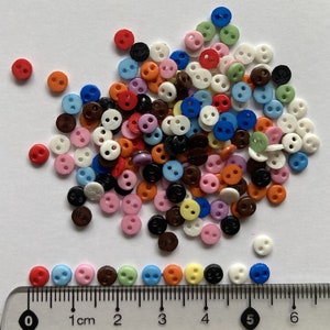 May include: A large assortment of colourful two-hole plastic buttons in various colours, including red, blue, green, yellow, white, black, brown, pink, and orange. The buttons are arranged in a pile on a white surface. A ruler is visible at the bottom of the image.