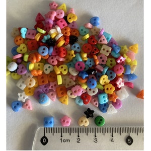 May include: A large assortment of colourful plastic buttons in various shapes, including hearts, stars, squares, and circles. The buttons are arranged on a white surface with a ruler measuring 5 centimetres.