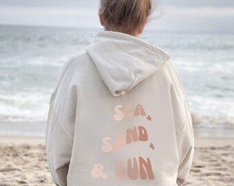 Puede incluir: Una sudadera con capucha blanca con el texto "SEA, SAND, & SUN" en rosa y naranja en la espalda. La sudadera la lleva puesta una persona de pie en una playa.