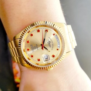 May include: A gold-tone wristwatch with a white face and red accents. The watch face features the text "West End Watch Co. Geneva" and "Prima Automatic 25 Jewels". The day of the week is displayed as "Tuesday 2".