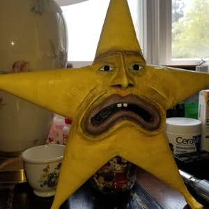 Puede incluir: Una escultura amarilla en forma de estrella con una cara humana. La cara tiene ojos marrones, una nariz marr&oacute;n y una boca marr&oacute;n con dientes. La escultura est&aacute; sentada en una mesa con un frasco de botones delante.