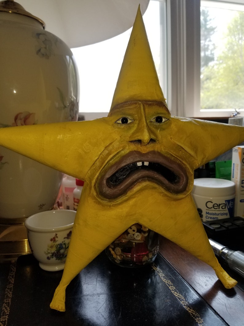 Puede incluir: Una escultura amarilla en forma de estrella con una cara humana. La cara tiene ojos marrones, una nariz marr&oacute;n y una boca marr&oacute;n con dientes. La escultura est&aacute; sentada en una mesa con un frasco de botones delante.