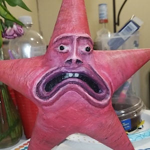 Puede incluir: Una escultura de cer&aacute;mica pintada a mano de una estrella de mar de dibujos animados rosa con una expresi&oacute;n de angustia y tristeza. La escultura tiene una superficie texturizada y est&aacute; pintada en tonos de rosa, morado y negro.