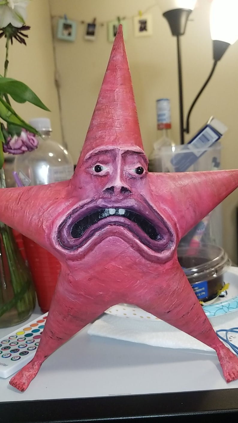 Puede incluir: Una escultura de cer&aacute;mica pintada a mano de una estrella de mar de dibujos animados rosa con una expresi&oacute;n de angustia y tristeza. La escultura tiene una superficie texturizada y est&aacute; pintada en tonos de rosa, morado y negro.