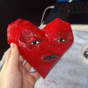 May include: A red paper mache heart with a painted face, including eyes, a nose, and a mouth. The heart is slightly misshapen and has a textured surface.