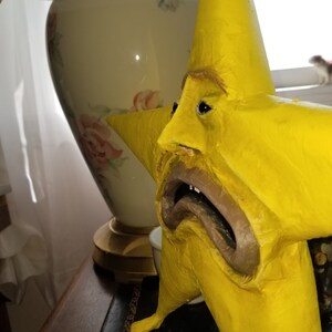 Puede incluir: Una estrella amarilla de papel mach&eacute; con una cara triste y una gran boca abierta. La estrella est&aacute; de pie sobre una superficie de madera.