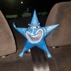 May include: A blue paper mache star with a smiling face and a pointed hat. The star is sitting in the back seat of a car.