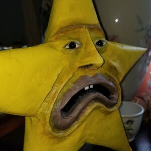 Puede incluir: Una escultura amarilla en forma de estrella con una cara humana. La cara tiene una expresi&oacute;n triste con la boca abierta y los dientes visibles. La escultura est&aacute; hecha de un material texturizado y est&aacute; pintada de un color amarillo brillante.