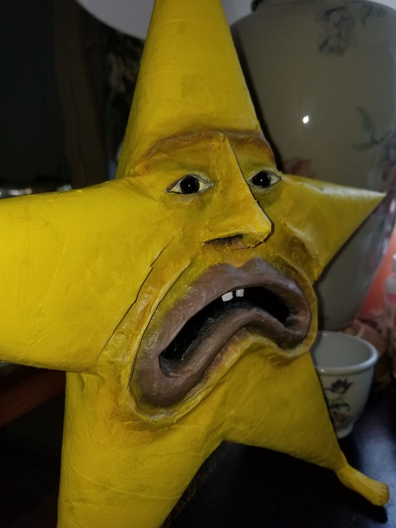 Puede incluir: Una escultura amarilla en forma de estrella con una cara humana. La cara tiene una expresi&oacute;n triste con la boca abierta y los dientes visibles. La escultura est&aacute; hecha de un material texturizado y est&aacute; pintada de un color amarillo brillante.