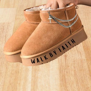 May include: A pair of tan suede ankle boots with thick platform soles. The boots feature a white faux fur lining and a silver-toned chain accessory with a cross charm attached to the side. The text WALK BY FAITH is printed on the platform sole.