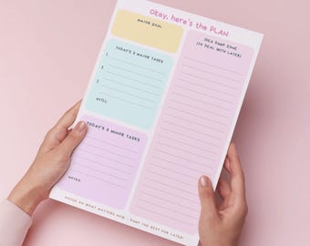 May include: A pastel-colored notepad with the text "Okay, here's the PLAN." The notepad is divided into sections for major goals, tasks, and notes. The notepad is held by a person.