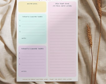 May include: A Daily Focus Planner notepad with a pastel color scheme. The notepad features sections for major goals, tasks, and an idea dump zone. Text includes "Daily Focus Planner", "Today's 3 Major Tasks", and "Today's 3 Minor Tasks". The notepad is on a beige fabric.