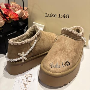 May include: A pair of brown suede ankle boots with a platform sole. The boots have a faux fur lining and are adorned with a pearl chain and a small bow. The text "Luke 1:45" is printed on the side of each boot.