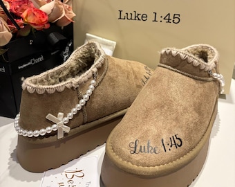 May include: A pair of brown suede ankle boots with a platform sole. The boots have a faux fur lining and are adorned with a pearl chain and a small bow. The text "Luke 1:45" is printed on the side of each boot.