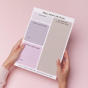 May include: A notepad with the text "Okay, here's the PLAN" in black. The notepad has sections for a major goal, tasks, and notes, with a large section for ideas. The notepad is held against a pink background.