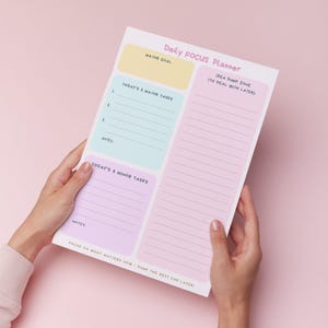 Może przedstawiać: Notatnik Daily Focus Planner z pastelowymi blokami kolorów na zadania i notatki. Notatnik zawiera sekcje na główne cele, zadania i strefę zrzutu pomysłów. Tekst na notatniku brzmi "Skup się na tym, co ważne teraz - resztę zostaw na później!"