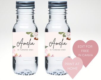 May include: Two clear plastic water bottles with pink floral labels that say "Amelia is turning one!"  Two pink heart-shaped stickers with the text "Edit for free in Canva" and "Print at home".