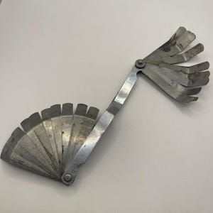 Può includere: Uno spessimetro in metallo argentato, a ventaglio, con più lame sottili di varie lunghezze. Le lame sono collegate da un punto di perno centrale, che consente di ripiegarle. Lo strumento è progettato per misurare fessure e giochi.