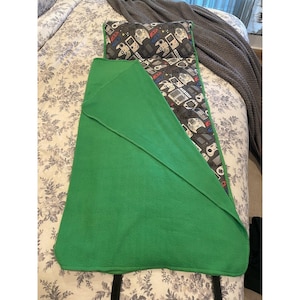 May include: A green sleeping bag with a matching pillow. The sleeping bag has a green exterior and a patterned interior with video game controllers in gray, white, red, and blue. The pillow has the same pattern. Black straps are attached to the bottom.