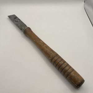 May include: A vintage wood chisel with a metal blade. The chisel has a long, wooden handle with carved rings near the end. The blade is rectangular and shows signs of wear.